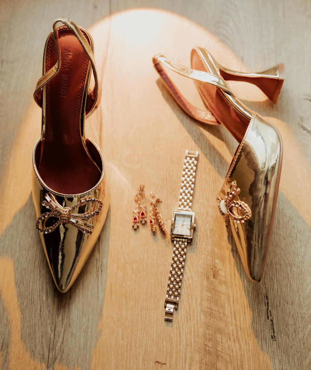 Gold heels, a wristwatch and earrings on a wooden surface